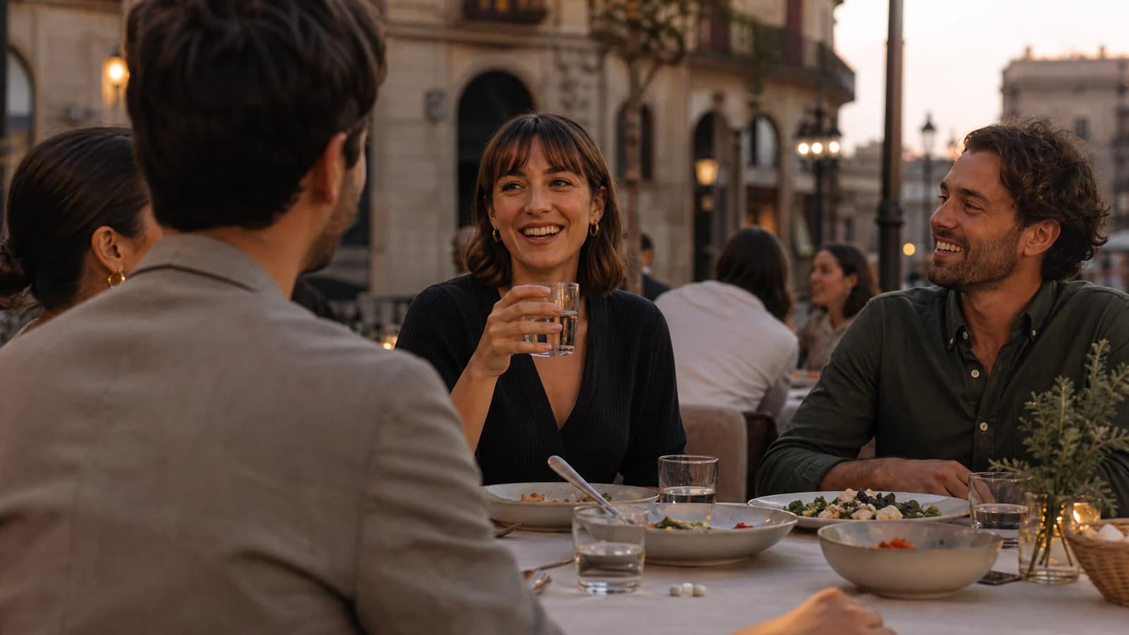 Amigos compartiendo cena y copas de agua en una terraza de un restaurante europeo al atardecer, con cápsulas en la mesa.