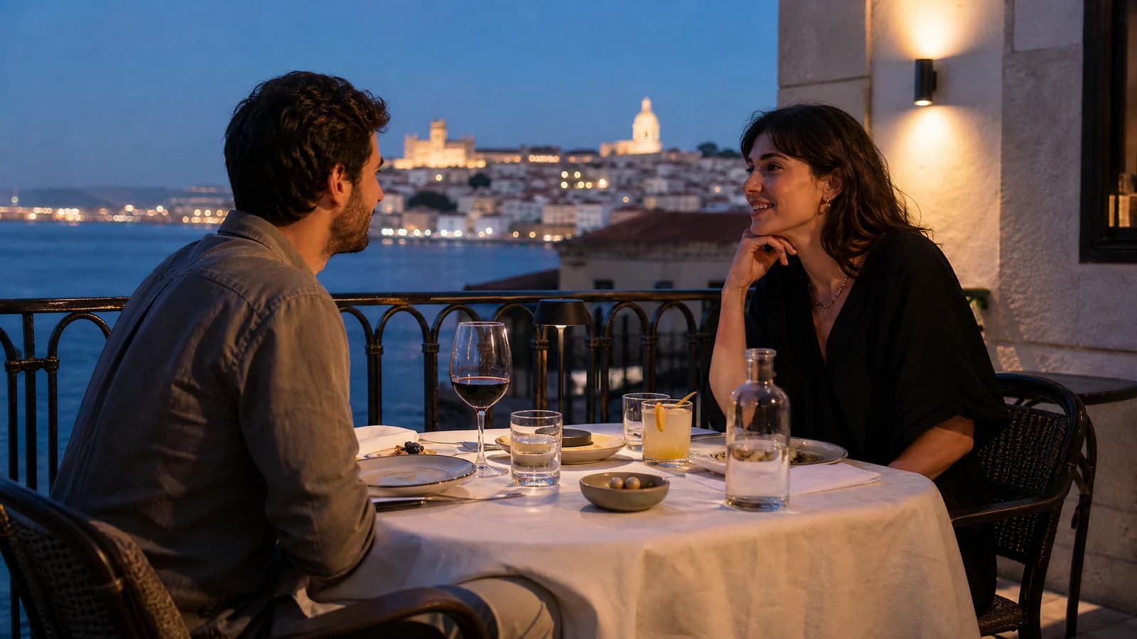 Un hombre y una mujer se sientan frente a frente en una mesa de restaurante en una terraza con vistas al mar, con bebidas y platos delante de ellos.